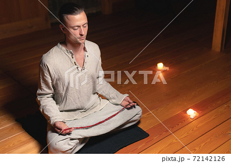 A man in clothes for practice and meditation sits in a lotus pose and holds red rosary to concentrate attention in a wooden room with dim light A man in clothes for practice and meditation sits in a lotus pose and holds red rosary to concentrate attention in a wooden room with dim light 72142126