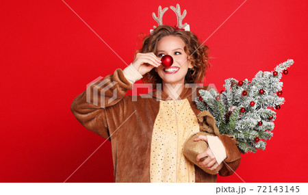 Cute young woman in a Christmas reindeer costume with red bauble and Christmas tree on colorful red background. Cute young woman in a Christmas reindeer costume with red bauble and Christmas tree on colorful red background. 72143145