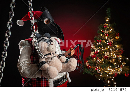 a toy Santa Claus in a plague doctor mask holds a bear dressed in a leather belt BDSM accessory against the background of a Christmas tree a toy Santa Claus in a plague doctor mask holds a bear dressed in a leather belt BDSM accessory against the background of a Christmas tree 72144675