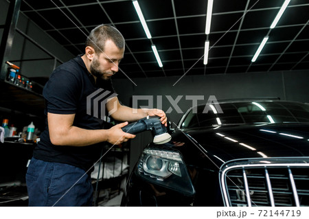 Young handsome Caucasian man, worker of auto detailing service, holds orbital polisher in the hand and polishes a hood of black modern car. Car detailing and polishing concept Young handsome Caucasian man, worker of auto detailing service, holds orbital polisher in the hand and polishes a hood of black modern car. Car detailing and polishing concept 72144719