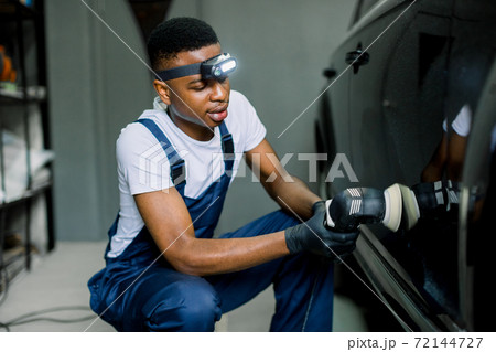 Auto detailing service, polishing of the car. Side view of young African American man worker in t-shirt and overalls, polishing black car body and door with orbital polisher and wax Auto detailing service, polishing of the car. Side view of young African American man worker in t-shirt and overalls, polishing black car body and door with orbital polisher and wax 72144727