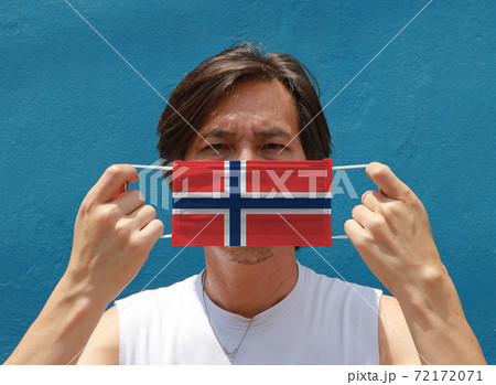 A man with Norway flag on hygienic mask in his hand and lifted up the front face on blue background. A man with Norway flag on hygienic mask in his hand and lifted up the front face on blue background. 72172071
