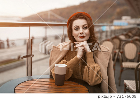 Lovely french young woman sitting at restaurant terrace with coffee mug looking at camera. Portrait of stylish young woman wearing autumn coat and red beret outdoors 72176609