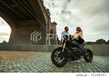 Young sexy woman hugging cute man in stylish black leather jacket, sitting on sports motorcycle under the bridge in the city on sunset and kissing 72189547