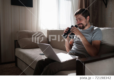 Funny man looking searching binoculars a laptop on the table working at home office. 72198342
