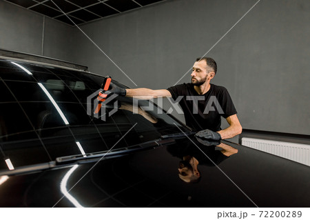 Anti rain coating at auto service. Male worker in black uniform and protective gloves, on a professional car wash service, applying anti rain coating on a windshield of luxury black car 72200289