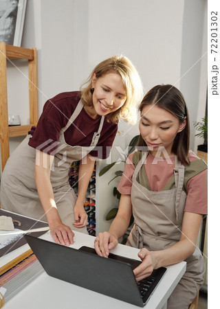 Happy young blond craftswoman in apron bending over colleague using laptop 72201302