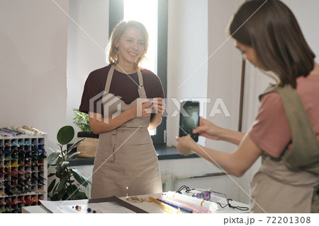 Happy young blond female with handmade glass bead standing by workplace Happy young blond female with handmade glass bead standing by workplace 72201308