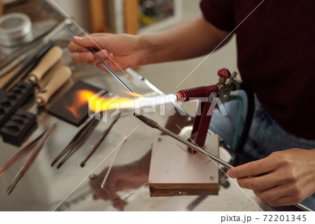 Hands of young female lampworker holding two workpieces over burner Hands of young female lampworker holding two workpieces over burner 72201345