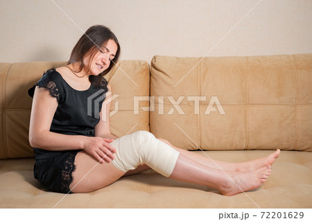 Crying barefoot lady with white elastic bandage on her knee sitting on brown sofa at home 72201629