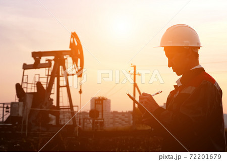 man in a white helmet stuffs paper with a pen against the background of an oil pump at sunset man in a white helmet stuffs paper with a pen against the background of an oil pump at sunset 72201679