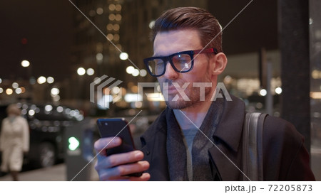 Man in eyeglasses with phone on background of business building in night time Man in eyeglasses with phone on background of business building in night time 72205873