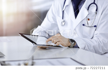 Unknown male doctor sitting and working with tablet computer iin a darkened clinic, glare of light on the background, close-up of hands 72227211