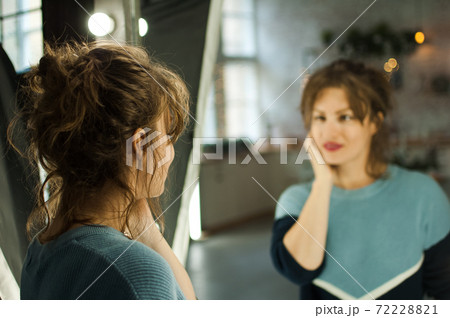 Young beautiful woman with perfect skin during winter season in blue sweater looking at the mirror at home in the room with Christmas decoration, Xmas tree and lights Young beautiful woman with perfect skin during winter season in blue sweater looking at the mirror at home in the room with Christmas decoration, Xmas tree and lights 72228821