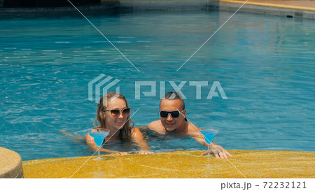 The loving couple hugs and kisses, drinking blue cocktail alcohol liquor in swimming pool at hotel outdoor. Portrait of caucasian man and woman. 72232121