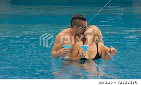 The loving couple hugs and kisses, drinking blue cocktail alcohol liquor in swimming pool at hotel outdoor. Portrait of caucasian man and woman. 72232130