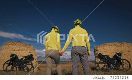 The man and woman travel on mixed terrain cycle touring with bikepacking. The two people journey with bicycle bags. Mountain snow capped, stone arch. The man and woman travel on mixed terrain cycle touring with bikepacking. The two people journey with bicycle bags. Mountain snow capped, stone arch. 72232218