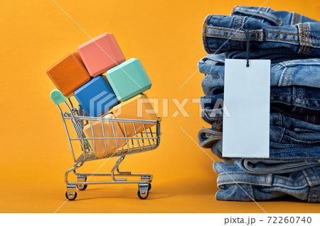 A stack of blue jeans with a white blank tag on a yellow background. Shopping trolley with multi-colored cubes. Sales consept 72260740