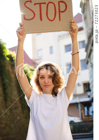 Young protesting woman in white shirt and jeans holds protest sign broadsheet placard with slogan 'Stop' for public demonstration on wall background. 72273023