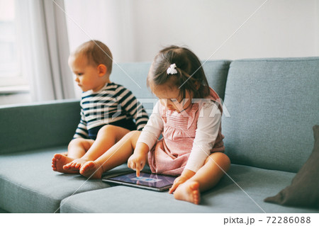 Two adorable babies are sitting on the couch and playing on a tablet. Brother and sister spend time playing together Two adorable babies are sitting on the couch and playing on a tablet. Brother and sister spend time playing together 72286088