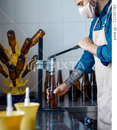 Beer packaging, brewery and factory work. Young attractive worker in apron and protective mask corks bottles Beer packaging, brewery and factory work. Young attractive worker in apron and protective mask corks bottles 72295783
