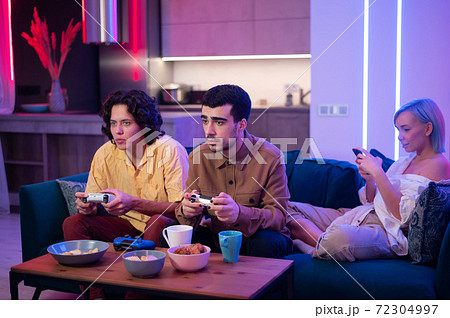 Guys playing video game while girl using smartphone for social media. Friends spending time together at home. Guys playing video game while girl using smartphone for social media. Friends spending time together at home. 72304997