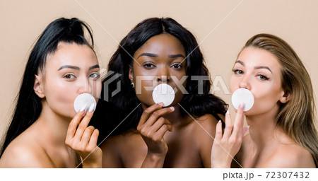 Skincare and natural beauty concept. Three diverse women with nude makeup, Asian, African and Caucasian, holding cotton pads, covering their lips, standing on beige background, close up 72307432