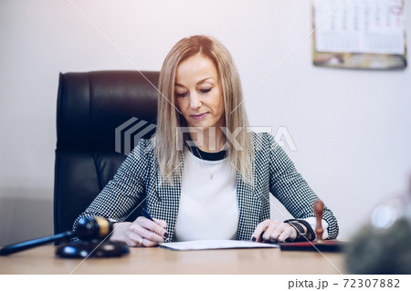 A female notary public sits at a wooden desk in her notary office signs documents. A female notary public sits at a wooden desk in her notary office signs documents. 72307882