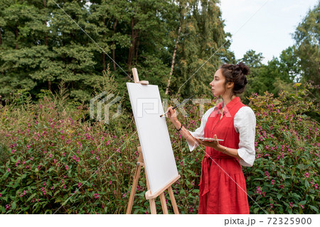 Getting started, woman in red dress, begins to paint picture, in summer on nature, background trees bushes flowers. Easel picture paint paintbrush. Free space for text. Inspiration and creativity. Getting started, woman in red dress, begins to paint picture, in summer on nature, background trees bushes flowers. Easel picture paint paintbrush. Free space for text. Inspiration and creativity. 72325900