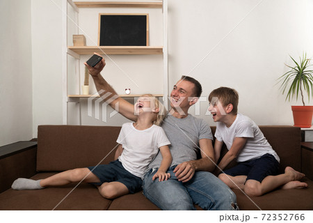 Young father and two sons take pictures of themselves on smartphone on the couch at home. Funny time Young father and two sons take pictures of themselves on smartphone on the couch at home. Funny time 72352766