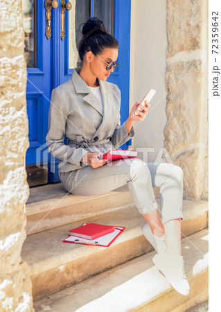 Woman in sunglasses is sitting and using smartphone near the door in the European city. 72359642