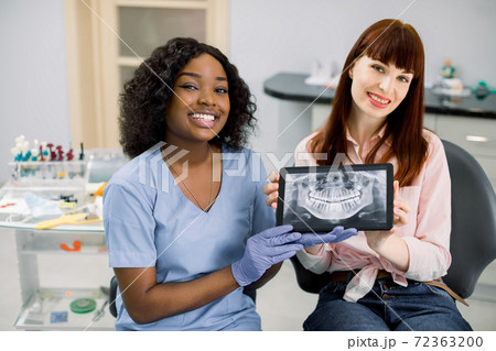 Shot of professional female african dentist and caucasian woman patient sitting in dentistry chair, showing digital tablet with patient oral scan, jaws and teeth x-ray, to camera and smiling 72363200