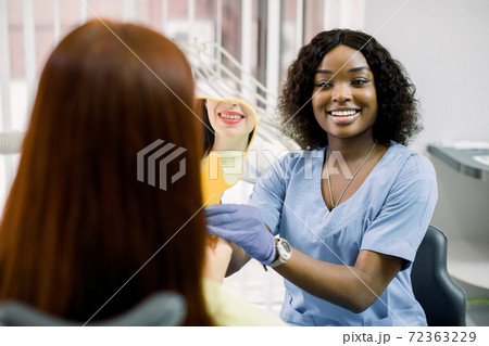 Back view of woman patient looking at her smile after curing or whitening teeth in dental clinic, and happy African female dentist, showing the mirror with reflection of smile Back view of woman patient looking at her smile after curing or whitening teeth in dental clinic, and happy African female dentist, showing the mirror with reflection of smile 72363229