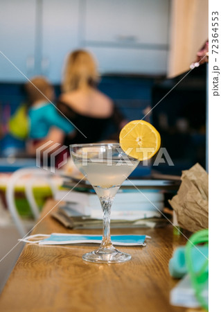 Home party in new normal, everyday life in lockdown, work life balance. Glass with cocktail on the kitchen table near laptop in home clutter and mother with baby on background Home party in new normal, everyday life in lockdown, work life balance. Glass with cocktail on the kitchen table near laptop in home clutter and mother with baby on background 72364553