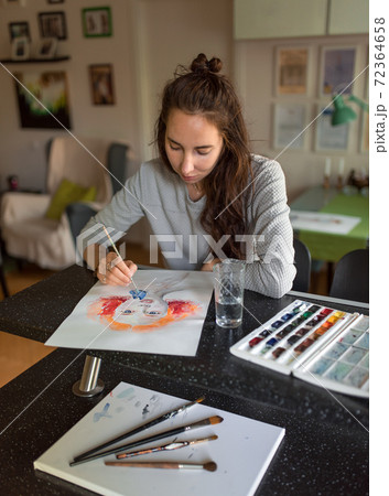 Woman at home at the table, draws paints, portrait of a girl. Bright orange paints, color palette. Black table, brush painting. Home studio creativity and art concept. Woman at home at the table, draws paints, portrait of a girl. Bright orange paints, color palette. Black table, brush painting. Home studio creativity and art concept. 72364658