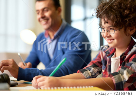 Portrait of smiling teenaged latin boy in glasses making notes while sitting at the desk together with his father and doing homework at home 72368463