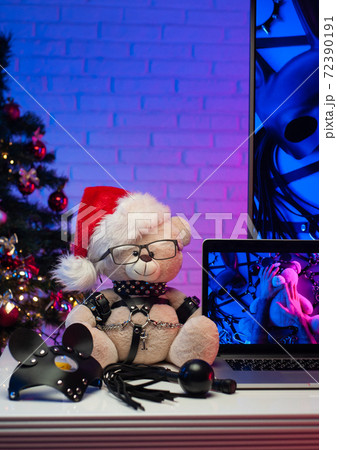 a Teddy bear in a leather belt an accessory for BDSM games in a Santa Claus hat next to a laptop a neon TV and a Christmas tree 72390191