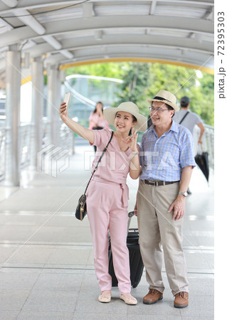 full length asian traveler couple husband and wife in pink and blue shirt with white nice hat and luggage walking in downtown with happy smiling face and taking photo from mobile while showing victory full length asian traveler couple husband and wife in pink and blue shirt with white nice hat and luggage walking in downtown with happy smiling face and taking photo from mobile while showing victory 72395303