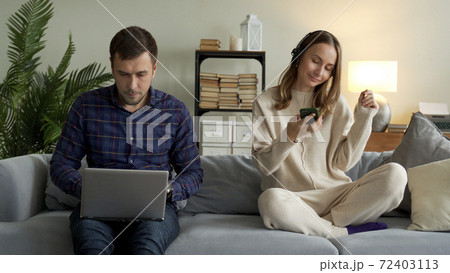 Happy couple with modern gadgets at home. A man is working on a laptop and a girl with headphones listening to music on a mobile phone is resting on the couch 72403113