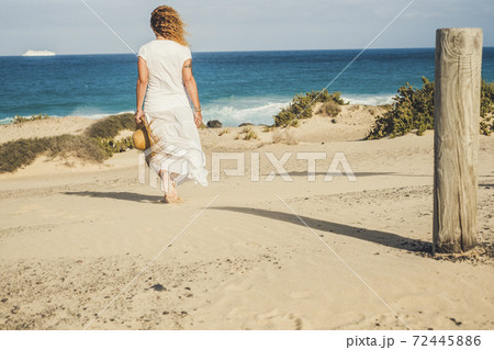 Caucasian woman with white dress viewed from back walking alone on the sand to the beach and blue ocean - water and sky in horizon background - vacation and travel concept 72445886