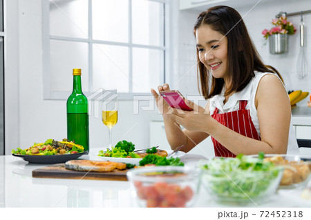 Asian woman in red apron taking a picture of food she make with mobile phone. Concept woman preparing meals at home. Asian woman in red apron taking a picture of food she make with mobile phone. Concept woman preparing meals at home. 72452318