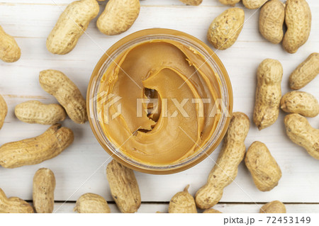 A jar of peanut butter on a white wooden table with peanuts scattered nearby. A jar of peanut butter on a white wooden table with peanuts scattered nearby. 72453149
