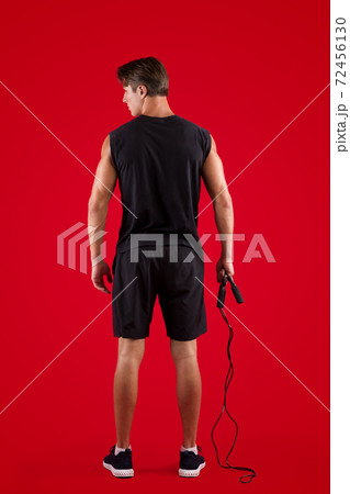 Aerobic workout. Back view of strong young man standing with skipping rope on red studio background Aerobic workout. Back view of strong young man standing with skipping rope on red studio background 72456130