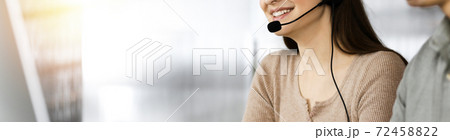Two smiling people in headsets are talking to the clients, while sitting at the desk in sunny modern office Two smiling people in headsets are talking to the clients, while sitting at the desk in sunny modern office 72458822