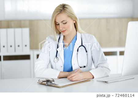 Woman-doctor at work while sitting at the desk in hospital or clinic. Blonde cheerful physician filling up medication history record form Woman-doctor at work while sitting at the desk in hospital or clinic. Blonde cheerful physician filling up medication history record form 72460114