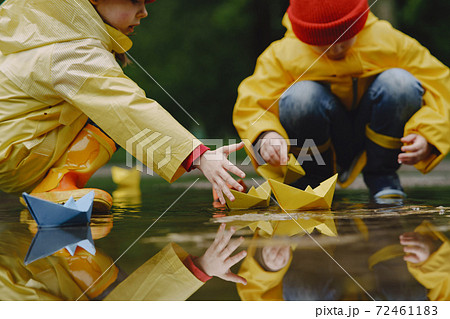 Funny kids in rain boots playing with paper ship by a puddle 72461183