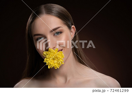 Smiling beauty model with bare shoulders holding a flower in her mouth with a beauty skin and long hair looking at camera isolated on black background. Woman nature cosmetic spa concept Smiling beauty model with bare shoulders holding a flower in her mouth with a beauty skin and long hair looking at camera isolated on black background. Woman nature cosmetic spa concept 72461299