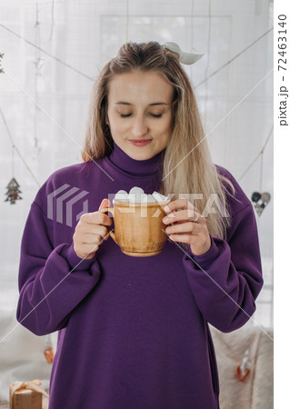Cozy winter Christmas mood. Young woman at home dress with coffee Cup and marshmallows in hand standing near xmas tree at home. Winter xmas weekends in home. Cozy homely concept Cozy winter Christmas mood. Young woman at home dress with coffee Cup and marshmallows in hand standing near xmas tree at home. Winter xmas weekends in home. Cozy homely concept 72463140