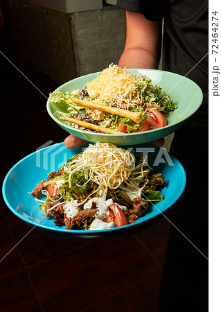 Waiter holds plates with warm salad with beef and smoked cheese and Caesar salad Waiter holds plates with warm salad with beef and smoked cheese and Caesar salad 72464274