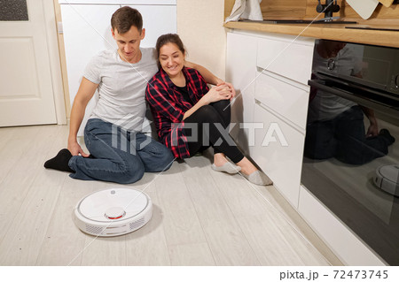 Young couple of man and woman watching robot vacuum cleaner while sitting on floor Young couple of man and woman watching robot vacuum cleaner while sitting on floor 72473745
